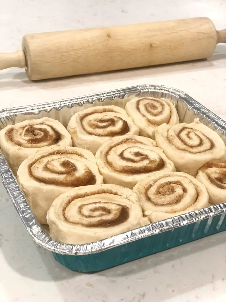 Nine cinnamon rolls rising in a pan on a counter with a rolling pin 