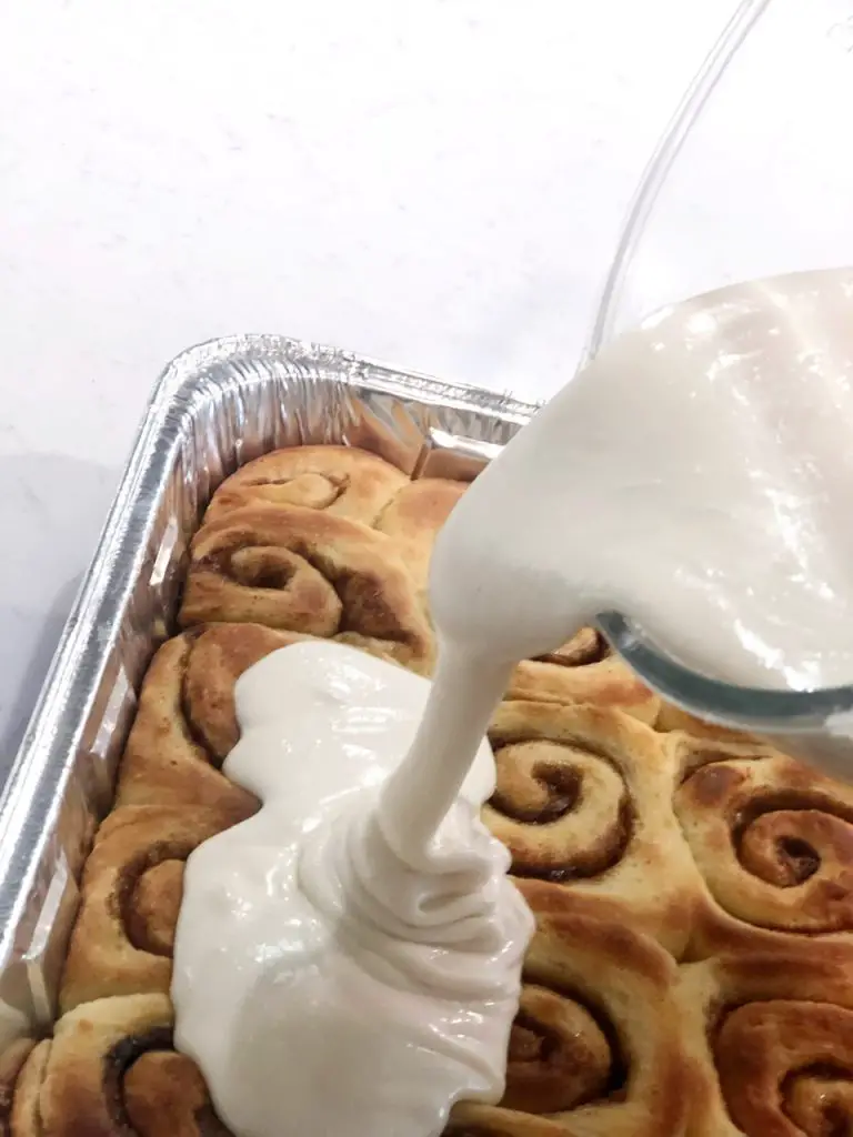 Pouring vanilla icing over a pan of baked cinnamon rolls