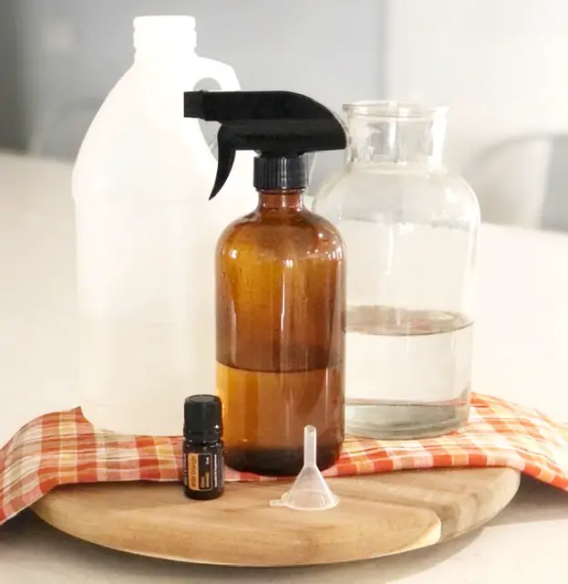 amber glass spray bottle, vinegar bottle, glass jar with water, small funnel and wild orange essential oil on wooden cutting board