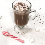 Glass of hot chocolate with cool whip and chocolate shavings on white counter with candy cane spoon
