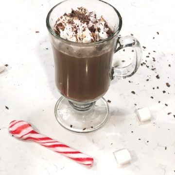 Glass of hot chocolate with cool whip and chocolate shavings on white counter with candy cane spoon