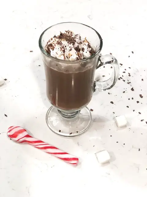 Glass of hot chocolate with cool whip and chocolate shavings on white counter with candy cane spoon