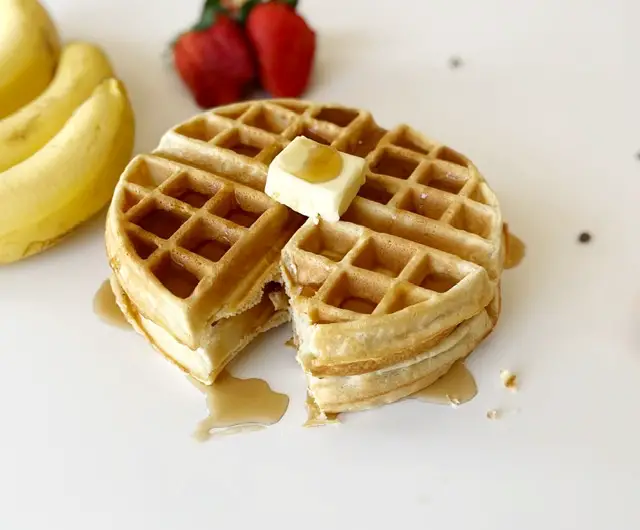 stacked gluten free waffles topped with butter and syrup sitting beside bananas and strawberries 