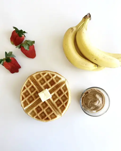 gluten free waffle topped with butter sitting beside bananas, strawberries and peanut butter