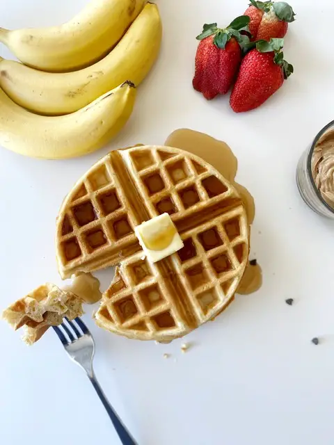 gluten free waffle topped with butter and syrup sitting beside bananas, strawberries and peanut butter