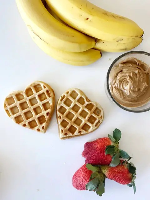 heart shaped gluten free waffles sitting beside peanut butter, bananas and strawberries 