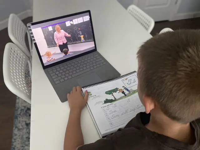 Boy doing abeka academy on computer with workbook on white table 