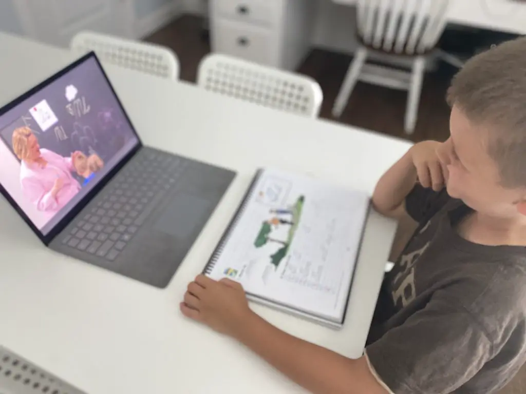 Boy doing abeka academy on computer with workbook on white table 