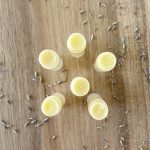 6 tubes of homemade beeswax lip balm in a circle with lavender petals on butcher block